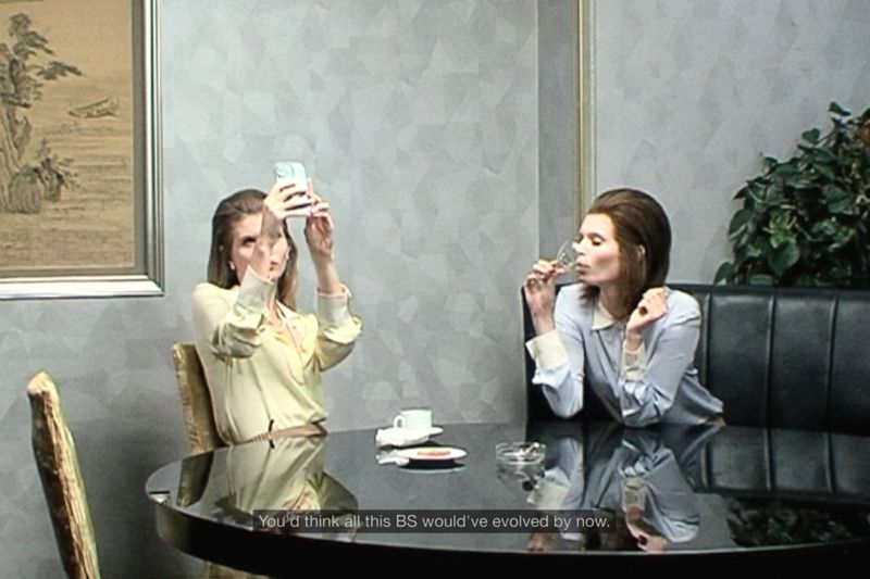 Two adult women seated at a round black table indoors; one is drinking from a glass, the other holds up a smartphone taking a photo.