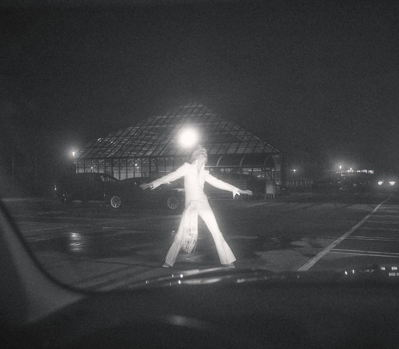 Adult in white jumpsuit stands in a parking lot at night, arms outstretched, with a glass building and cars visible in the background.