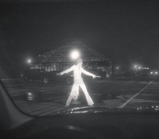 Adult in white jumpsuit stands in a parking lot at night, arms outstretched, with a glass building and cars visible in the background.
