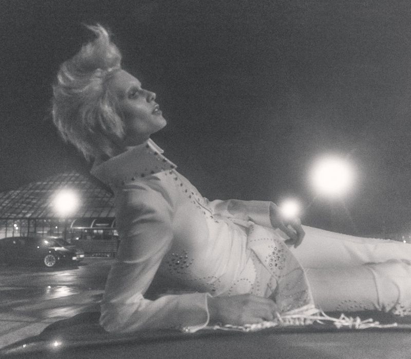 Monochrome image of a person with a prominent hairstyle reclining in a white embellished jumpsuit under dim light with vehicles in the background.