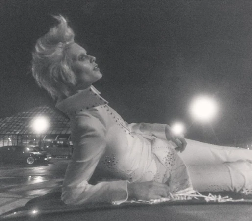 Monochrome image of a person with a prominent hairstyle reclining in a white embellished jumpsuit under dim light with vehicles in the background.