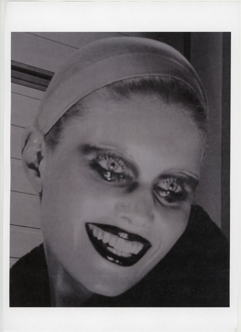 Close-up black-and-white portrait of a smiling adult woman with exaggerated makeup, wearing a head covering against a neutral backdrop.