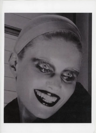 Close-up black-and-white portrait of a smiling adult woman with exaggerated makeup, wearing a head covering against a neutral backdrop.