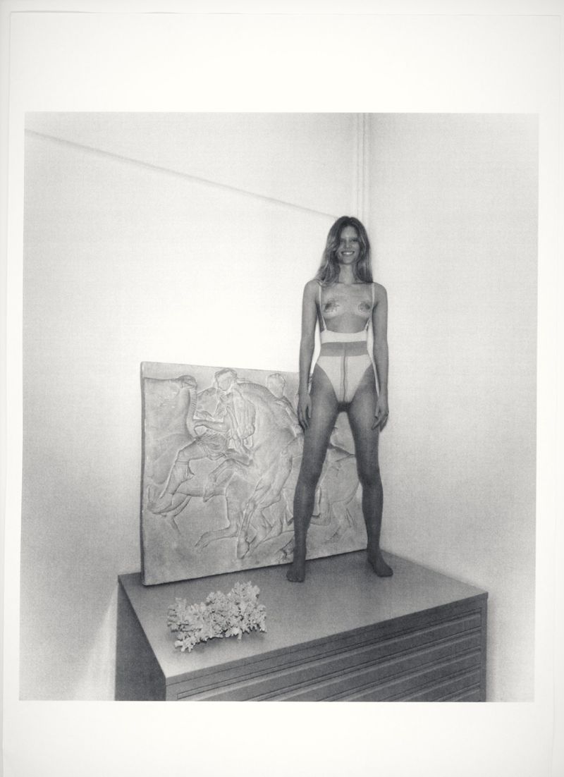 Artistic monochrome photo of a woman in tights standing on a stacked surface next to a bas-relief and a small bunch of flowers.