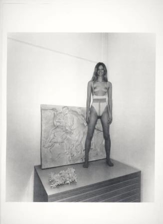 Artistic monochrome photo of a woman in tights standing on a stacked surface next to a bas-relief and a small bunch of flowers.