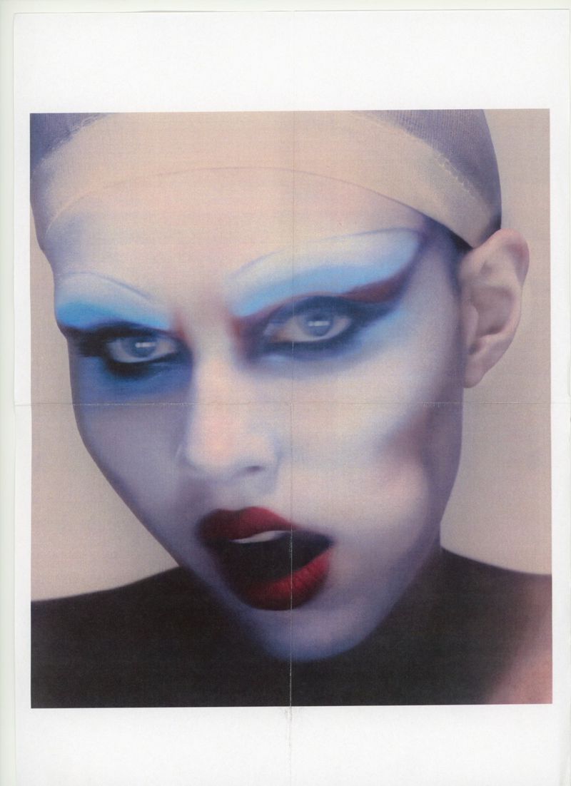 Headshot of a woman with dramatic blue and red makeup, a neutral head covering, and red lipstick, set against a plain, off-white background.