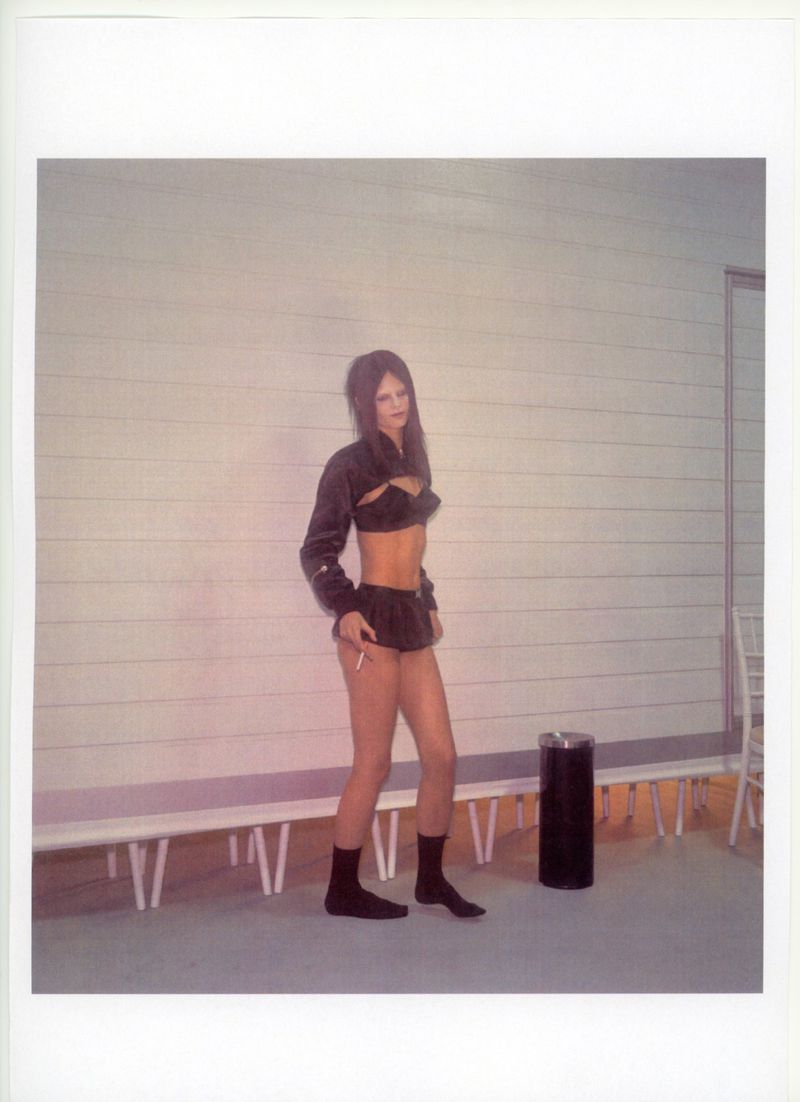 A woman in a black two-piece outfit stands next to a trash can in an indoor location with a bench and a white wall backdrop.