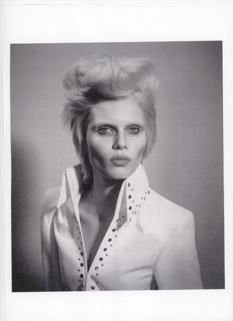 Monochrome portrait of adult woman with blonde hair styled in a high pompadour, wearing white collared shirt with studded trim. 
