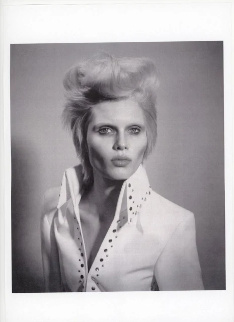 Monochrome portrait of adult woman with blonde hair styled in a high pompadour, wearing white collared shirt with studded trim. 