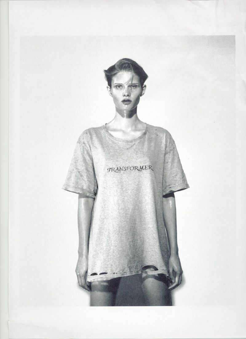 Monochrome studio portrait of a female model wearing a t-shirt with text, styled with slicked-back hair, against a light backdrop.