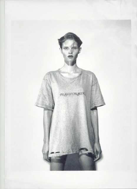 Monochrome studio portrait of a female model wearing a t-shirt with text, styled with slicked-back hair, against a light backdrop.
