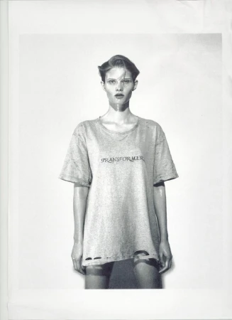 Monochrome studio portrait of a female model wearing a t-shirt with text, styled with slicked-back hair, against a light backdrop.