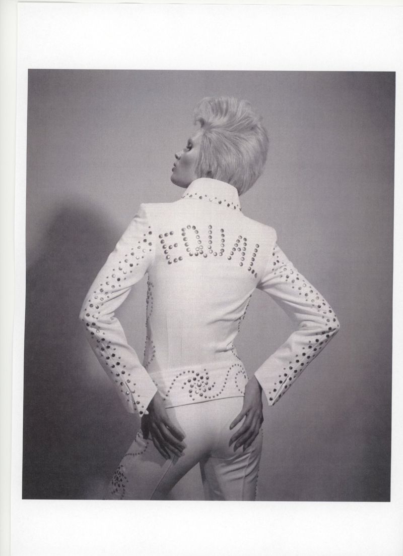 Blonde woman in white studded suit with 'EQUAL' on back turns away from camera in photo studio on grey background. Fashionable, bold style.