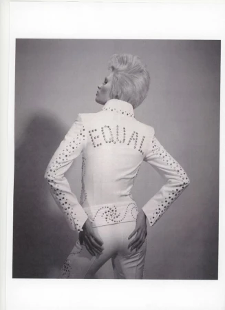 Blonde woman in white studded suit with 'EQUAL' on back turns away from camera in photo studio on grey background. Fashionable, bold style.