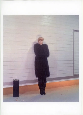 A woman in a dark coat stands against a horizontal-striped wall in an underground location with a black trashcan and orange accent stripe.