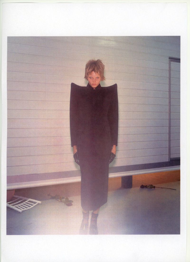 Woman with blonde hair and oversized-shoulder black dress stands in front of a paneled wall in a fashion photography portrait.