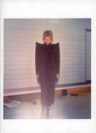 Woman with blonde hair and oversized-shoulder black dress stands in front of a paneled wall in a fashion photography portrait.