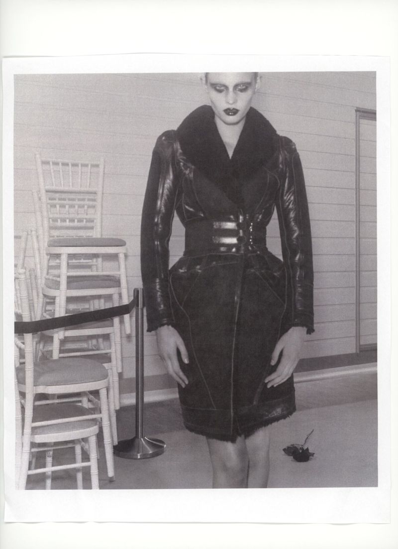 A woman poses in a black fur-collared coat, cinched at the waist with chairs and a chrome pole in the background in a studio.