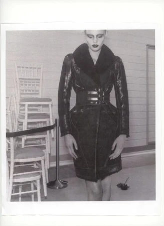 A woman poses in a black fur-collared coat, cinched at the waist with chairs and a chrome pole in the background in a studio.