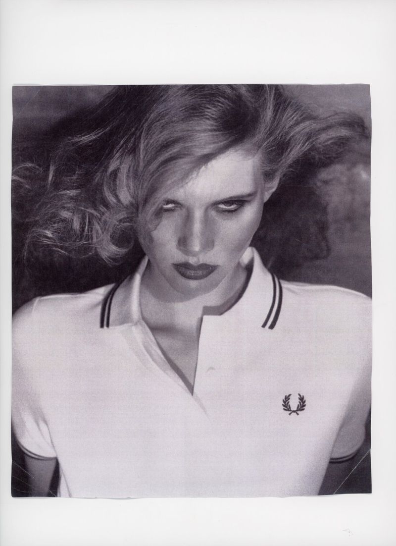 Monochrome portrait of a woman with wavy hair wearing a white polo shirt and dark lipstick, gazing intensely at the viewer.