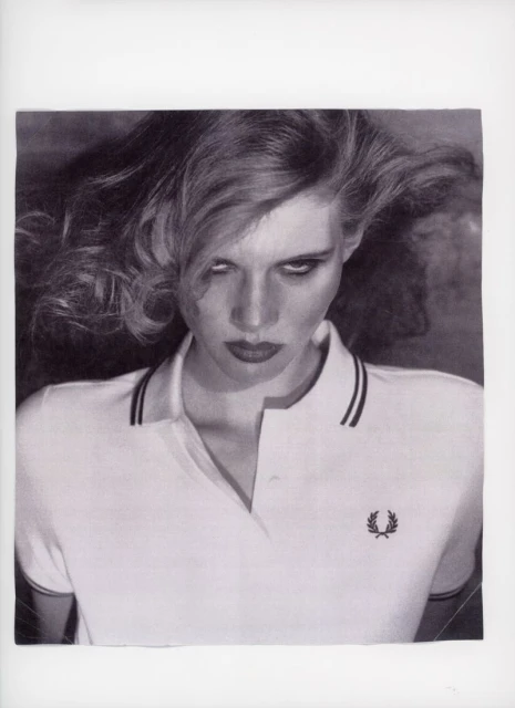 Monochrome portrait of a woman with wavy hair wearing a white polo shirt and dark lipstick, gazing intensely at the viewer.