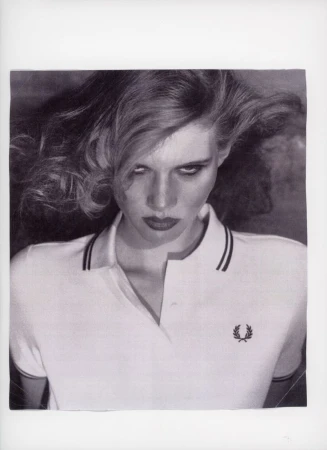 Monochrome portrait of a woman with wavy hair wearing a white polo shirt and dark lipstick, gazing intensely at the viewer.