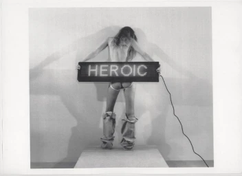 Model standing with back to camera holding an illuminated LED sign reading HEROIC against a white seamless background