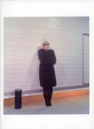 Model Ida Heiner posing in a voluminous black fur coat and dark boots against a horizontal paneled wall beside an ashcan.