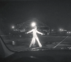 Monochrome photo of model Ida Heiner in a white flared jumpsuit posing in a dark parking lot with a greenhouse in the background.