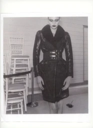 Model Ida Heiner wearing a structured black belted shearling coat next to stacked white chairs in monochrome.