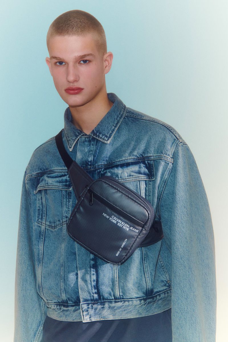 Close-up of a young adult male fashion model wearing a denim jacket, with a Calvin Klein crossbody bag in a studio setting.