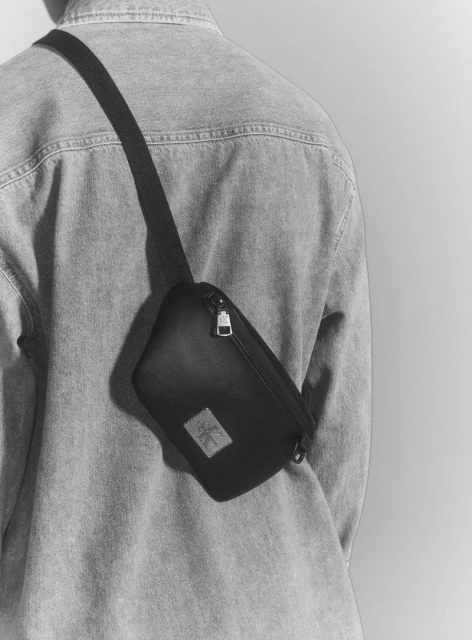 Black fanny pack with a strap worn by a person in a denim shirt against a neutral background, studio shot, fashion product photography.