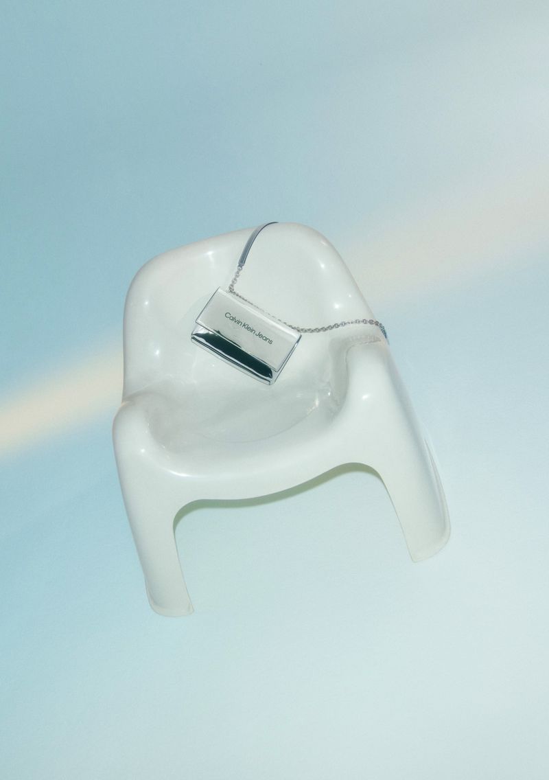 Calvin Klein Jeans purse on a white plastic chair against a light blue backdrop with soft, diffused lighting for a modern look.