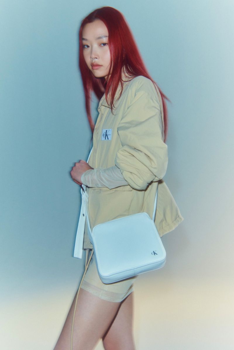 A stylish woman with auburn hair poses indoors, wearing a beige jacket, matching shorts, and a light blue Calvin Klein crossbody bag.
