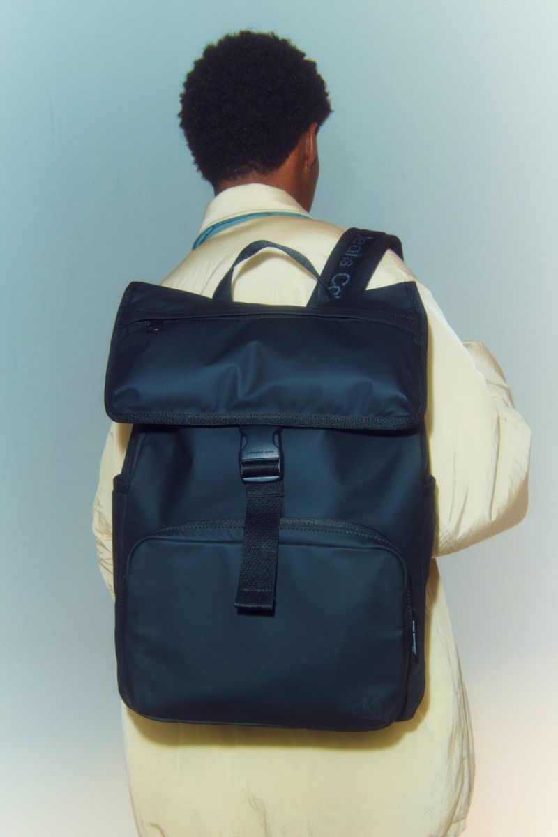 Person with dark hair wears a black backpack and beige jacket against a soft blue background in a studio product shot.