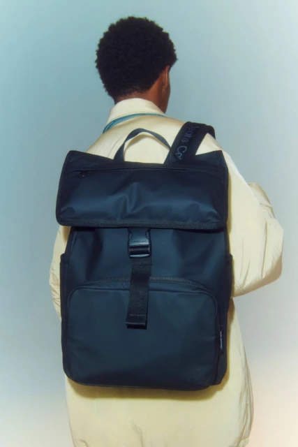 Person with dark hair wears a black backpack and beige jacket against a soft blue background in a studio product shot.