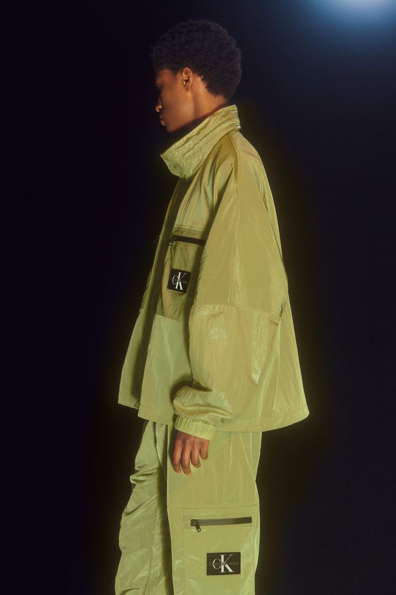 A person wearing a Calvin Klein olive-green jacket and pants stands indoors against a black backdrop and a bright, soft light.