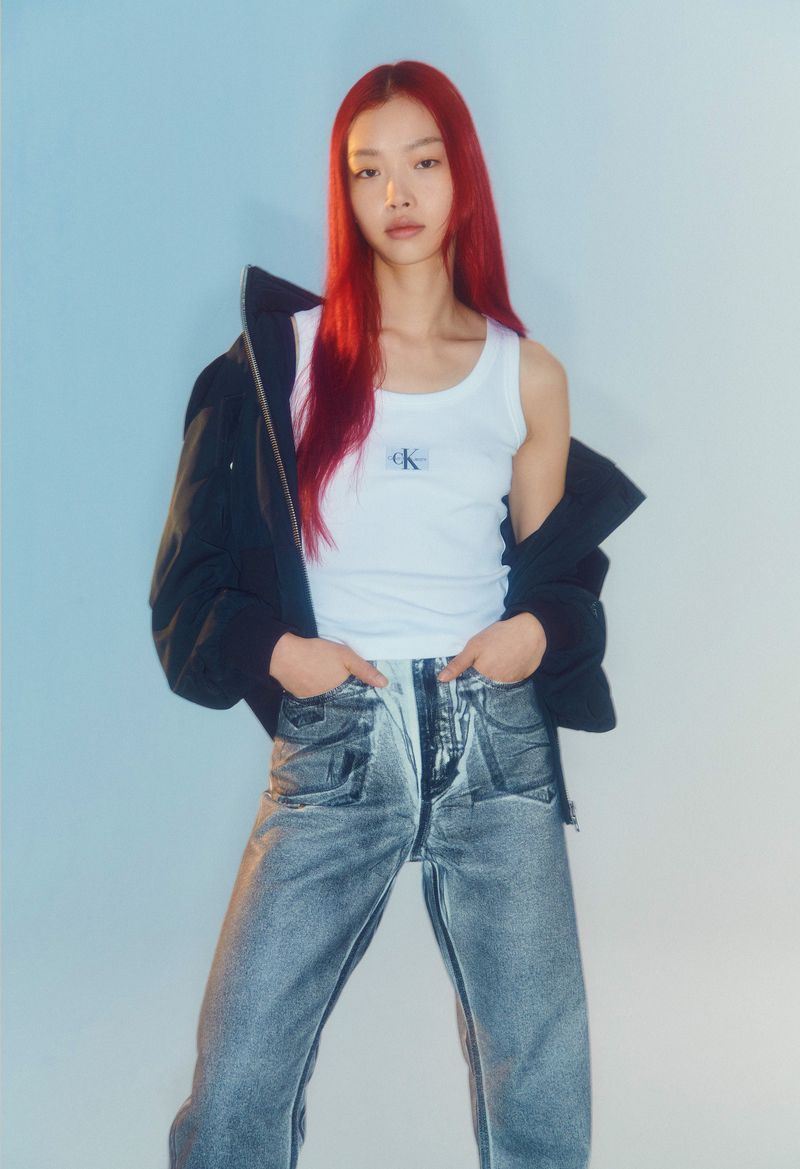 A slender Asian woman with red hair poses in a white tank top, acid-wash jeans, and a black jacket against a blue backdrop.