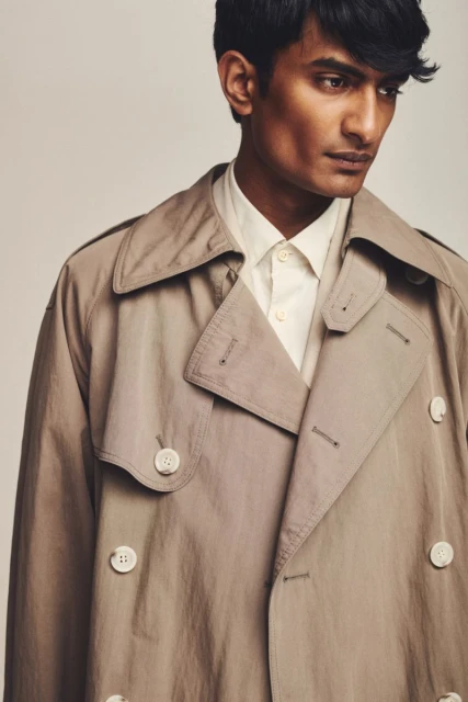 Close-up studio shot of a dark-haired male model wearing a khaki trench coat and a cream-colored collared shirt against a neutral backdrop.