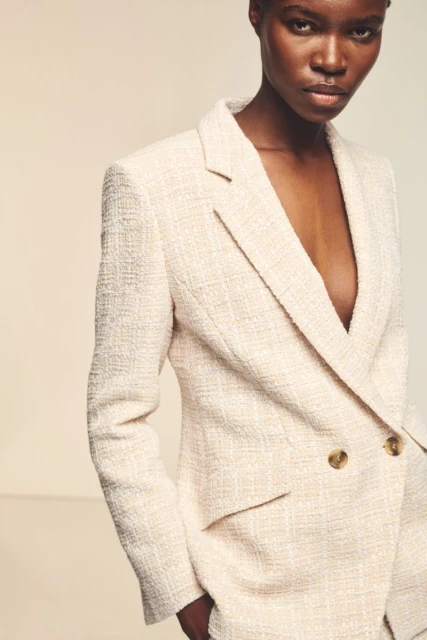 A woman with short hair poses in a double-breasted tweed blazer against a neutral background in a fashion editorial shot.