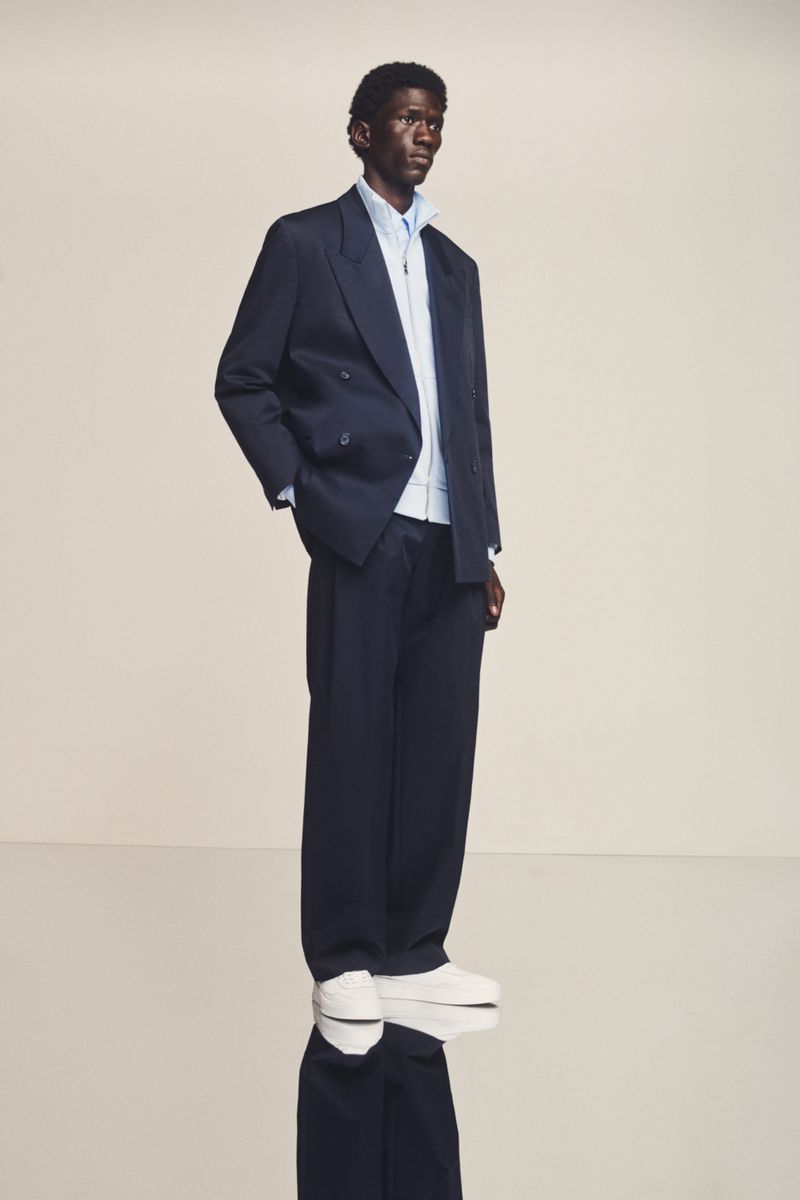A dark-skinned male model poses in a navy blazer and wide-leg trousers, layered over a light blue shirt and white trainers, indoor setting.