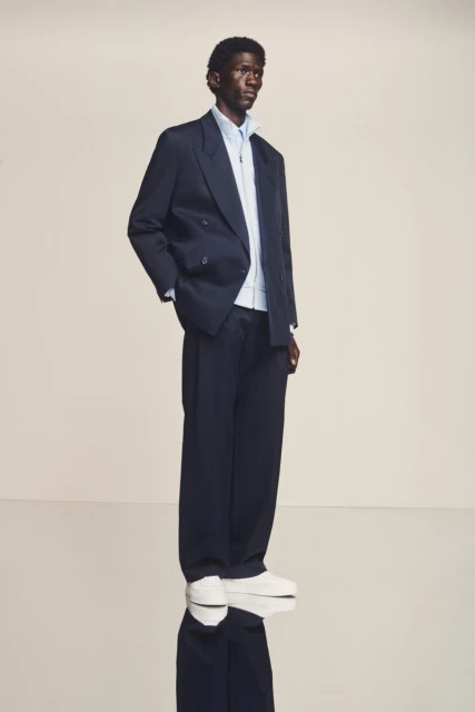A dark-skinned male model poses in a navy blazer and wide-leg trousers, layered over a light blue shirt and white trainers, indoor setting.