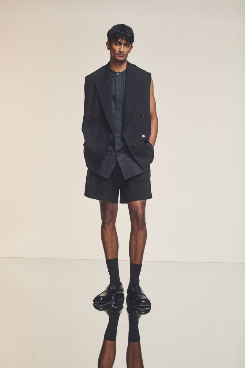 A man in a black blazer and shorts poses indoors with a neutral backdrop, showcasing a modern, high-fashion aesthetic and cool, sophisticated style.