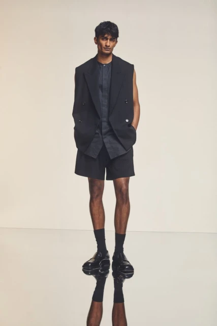 A man in a black blazer and shorts poses indoors with a neutral backdrop, showcasing a modern, high-fashion aesthetic and cool, sophisticated style.