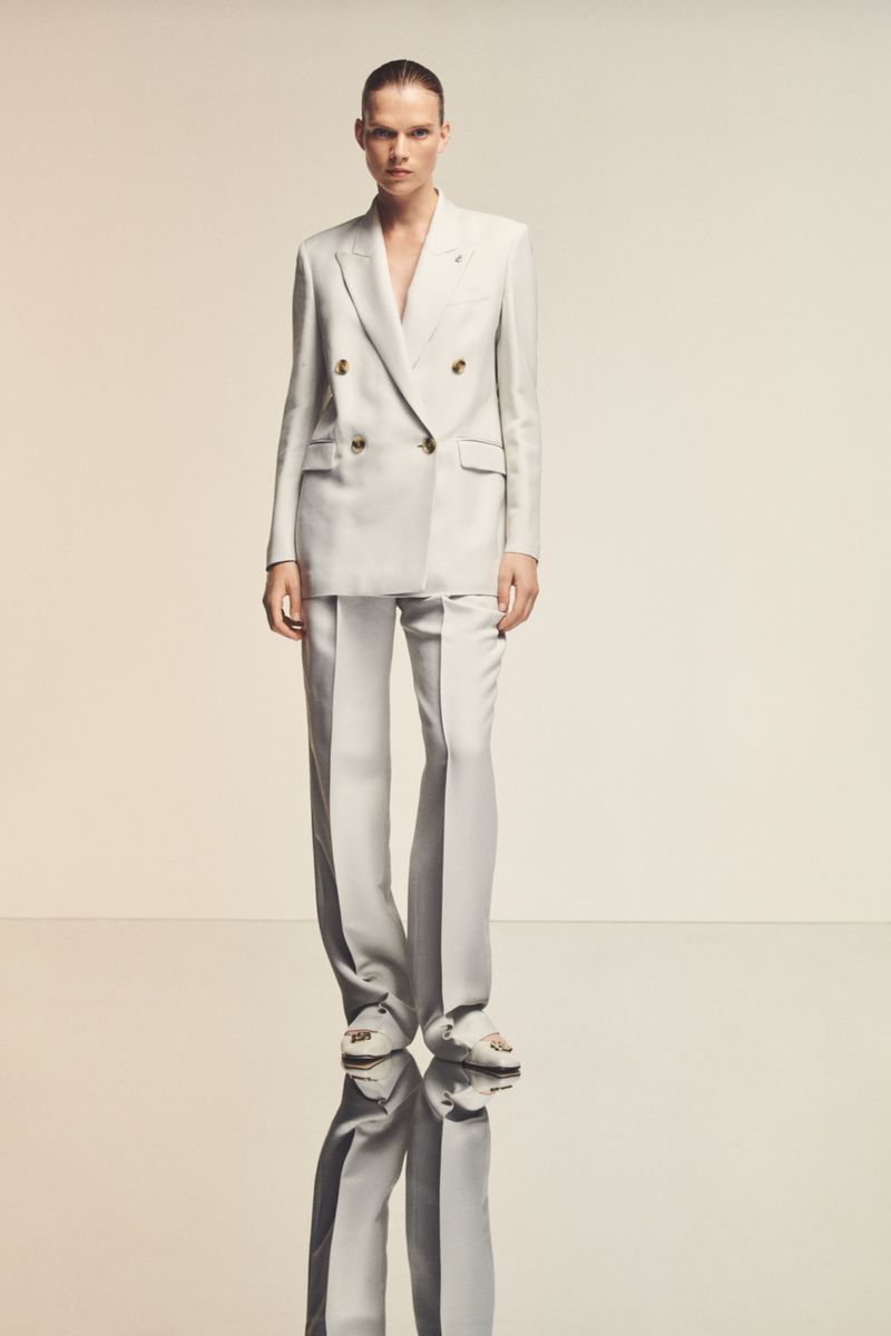 A woman poses in a white double-breasted suit with wide-leg trousers, standing on a reflective surface in a fashion editorial style.