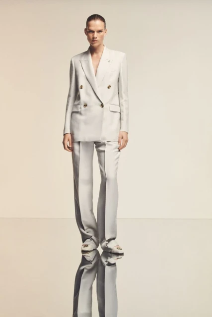 A woman poses in a white double-breasted suit with wide-leg trousers, standing on a reflective surface in a fashion editorial style.