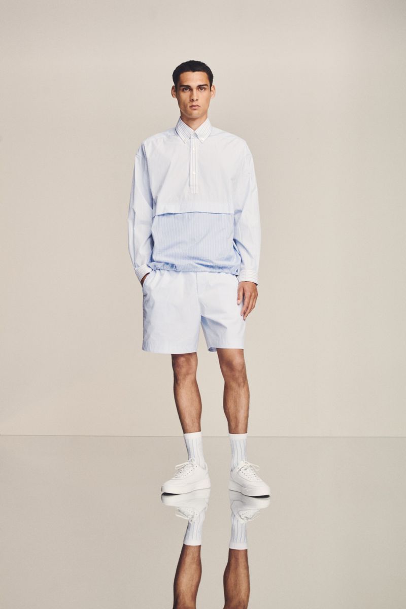 A full-length studio shot of a male fashion model in a white collared long-sleeve top with blue accents, white shorts, socks, and sneakers.