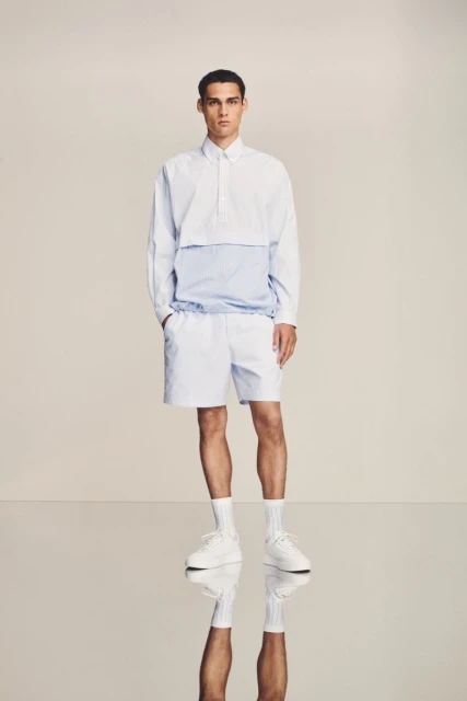 A full-length studio shot of a male fashion model in a white collared long-sleeve top with blue accents, white shorts, socks, and sneakers.