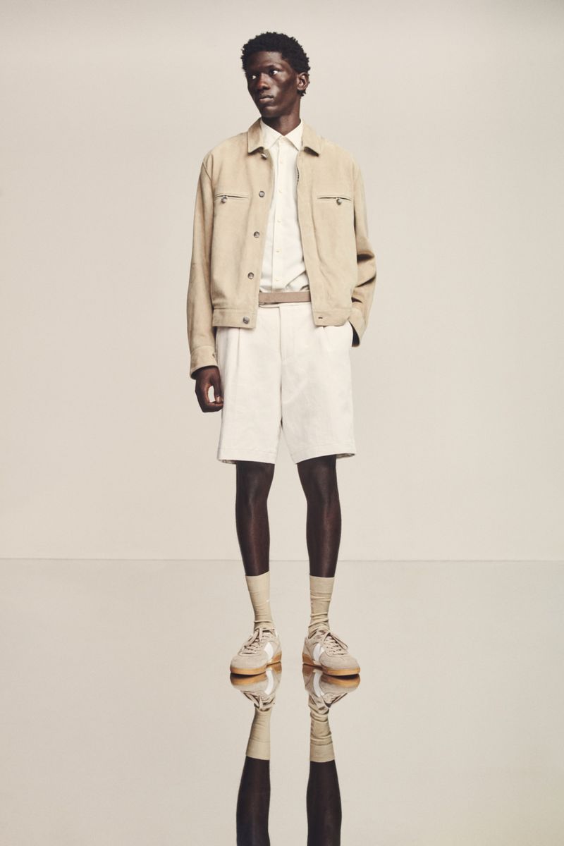 A full shot of a dark-skinned male model in light casual clothing, including a jacket, shorts, button-down shirt, socks, and shoes, posing in a studio.