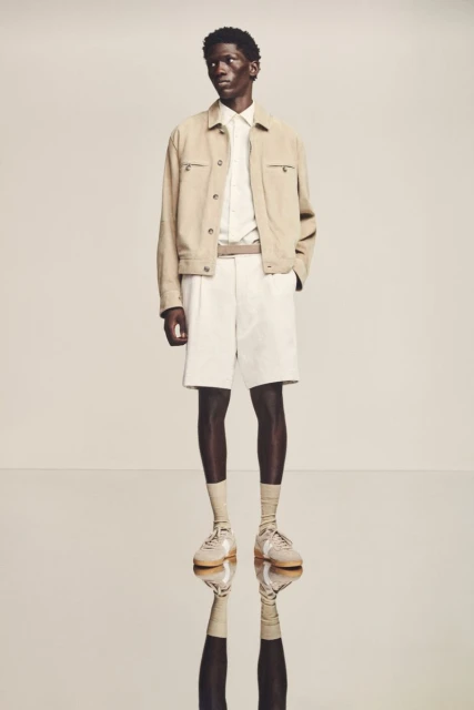 A full shot of a dark-skinned male model in light casual clothing, including a jacket, shorts, button-down shirt, socks, and shoes, posing in a studio.